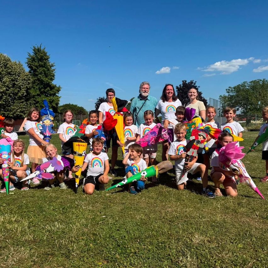 A group of children and adults are posing for a photo outdoors, all holding colorful kites. The children are wearing matching shirts and holding kites. Behind them, trees and a fence are visible.