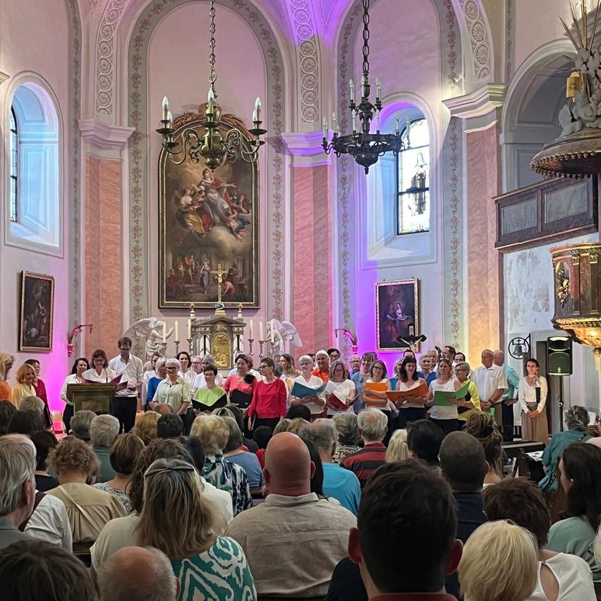 Ein Chor singt in einer Kirche mit rosa Wand, geschmückt mit Gemälden und Kronleuchtern. Das Publikum, Bücher in der Hand, hört aufmerksam zu.