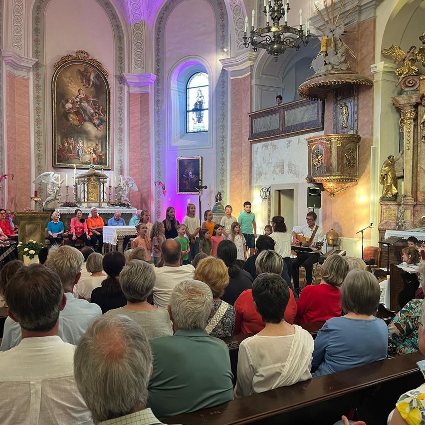 Ein Chor singt in einer Kirche mit einer Menschenmenge im Vordergrund. Im Hintergrund befindet sich eine Bühne mit einem Mikrofon und einem kerzenbeleuchteten Altar.