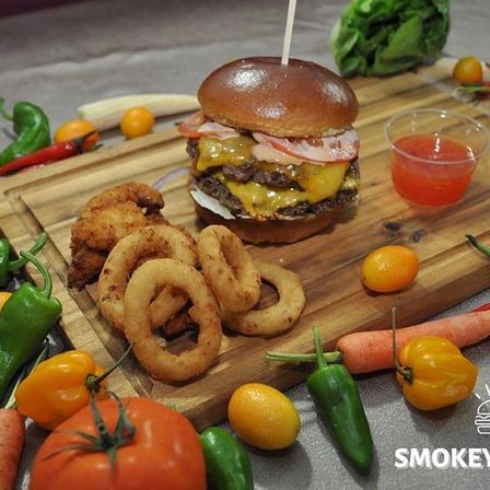 Ein Holzbrett zeigt einen Burger mit Käse, Fleisch und einer Tomatenscheibe. Gebratene Zwiebelringe und Hähnchenstücke sind ebenfalls auf dem Brett. Verschiedene Gemüsesorten, einschließlich Karotten, Paprika und Tomaten, umgeben das Brett.