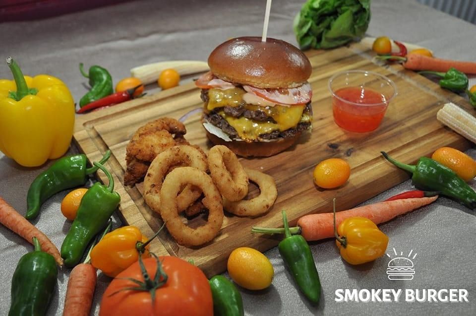 Ein Holzbrett zeigt einen Burger mit Käse, Fleisch und einer Tomatenscheibe. Gebratene Zwiebelringe und Hähnchenstücke sind ebenfalls auf dem Brett. Verschiedene Gemüsesorten, einschließlich Karotten, Paprika und Tomaten, umgeben das Brett.