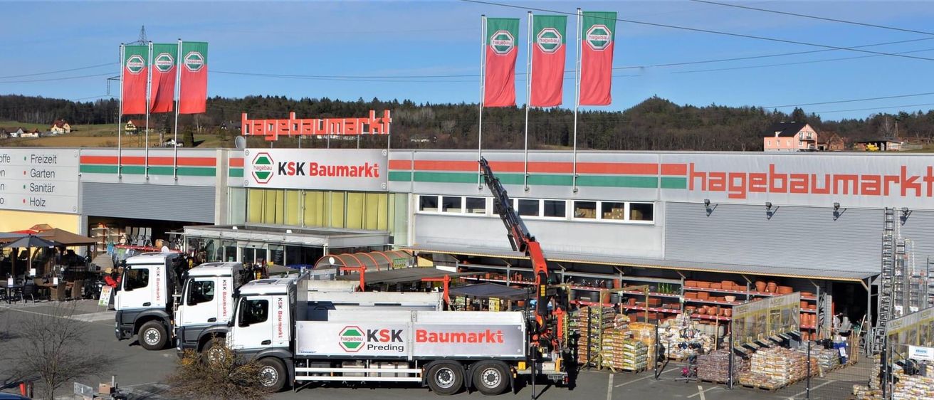 Ein KSK Baumarkt-Gebäude mit drei Flaggen darüber. Ein Lastwagen ist davor geparkt und ein Kran lädt etwas.