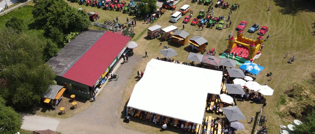Luftaufnahme einer großen Versammlung mit vielen Autos und Traktoren, einem Zelt und aufblasbaren Spielgeräten. Mehrere Gebäude und ein Feld mit Solarpanelen.