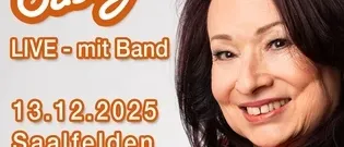 Eine lächelnde Frau mit schwarzen Haaren und einem orangefarbenen Oberteil ist in einer Werbung für Sassy LIVE mit Band am 13. Dezember 2025 in Saalfelden Congress zu sehen.