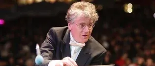 Ein Dirigent in einem schwarzen Anzug und Fliege hält einen Taktstock in einer Konzerthalle mit einem unscharfen Publikum im Hintergrund.