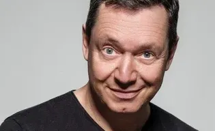 Ein lächelnder Mann mit kurzen Haaren, der ein schwarzes T-Shirt trägt, posiert vor einem grauen Hintergrund.