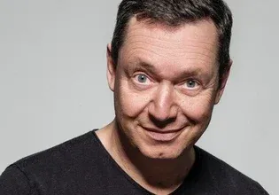 Ein lächelnder Mann mit kurzen Haaren, der ein schwarzes T-Shirt trägt, posiert vor einem grauen Hintergrund.