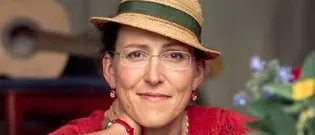 Eine lächelnde Frau in einem roten Kleid und einem Strohhut mit einem grünen Band lehnt sich gegen ein Blumenarrangement. Eine Gitarre ist hinter ihr.