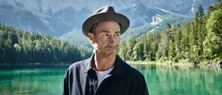 Ein Mann steht am Ufer eines ruhigen Sees, umgeben von üppigem Grün und hohen Bergen unter einem klaren blauen Himmel.