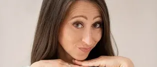 Eine Frau mit langen braunen Haaren, die ein blaues Oberteil trägt, macht einen komischen Gesichtsausdruck, wobei ihre Hände ein Dreieck am Kinn bilden, möglicherweise für ein Foto posierend.