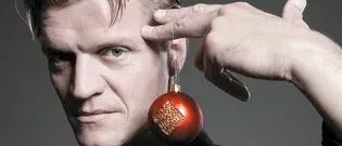 Ein Mann in einem schwarzen Hemd hält eine rote Weihnachtskugel mit goldenen Designs. Er starrt mit ernster Miene auf die Kugel.