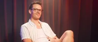 Ein Mann mit Brille sitzt auf einem Stuhl mit verschränkten Beinen. Er lächelt und schaut direkt in die Kamera. Er trägt ein weißes Poloshirt. Hinter ihm ist ein roter Vorhang mit einer Lampe darüber.