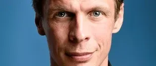 Ein Nahporträt eines Mannes mit ernster Miene, der ein schwarzes Hemd trägt und vor einem festen blauen Hintergrund steht.