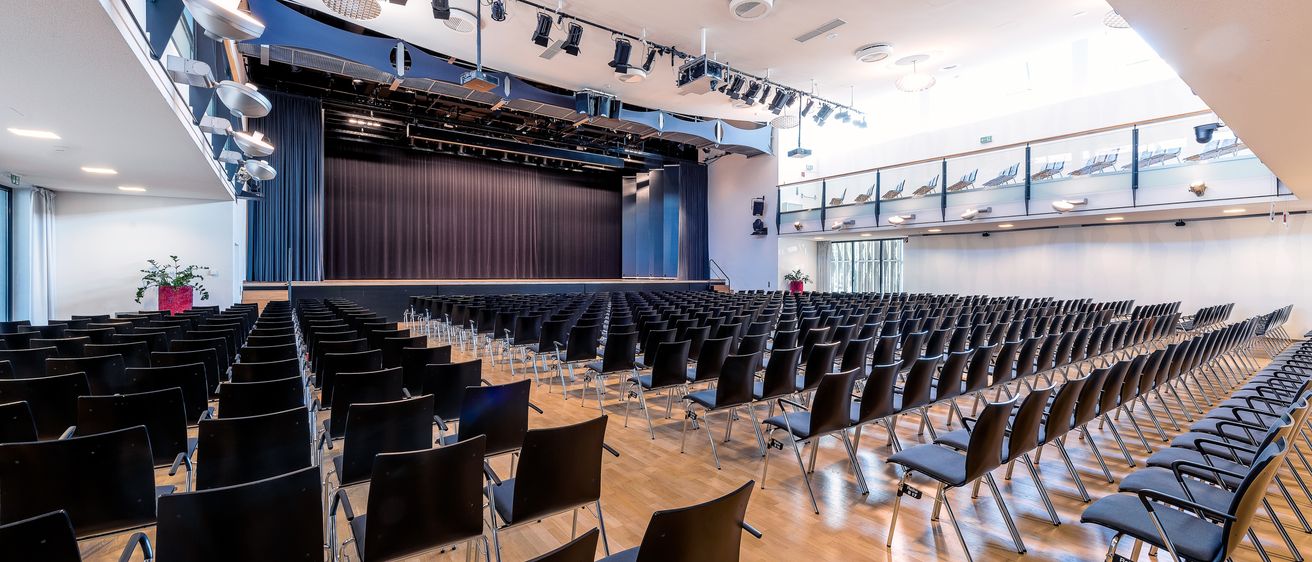 Ein leerer Auditorium mit Holzböden und Reihen schwarzer Stühle. Eine Bühne mit einem blauen Vorhang befindet sich vorne. Scheinwerfer und Lautsprecher sind an der Decke montiert.