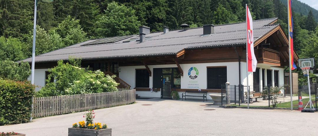 Ein Gebäude mit grauem Dach und Solarpanelen, umgeben von Bäumen und einem Zaun, mit Fahnen und einem Basketballkorb im Vordergrund.