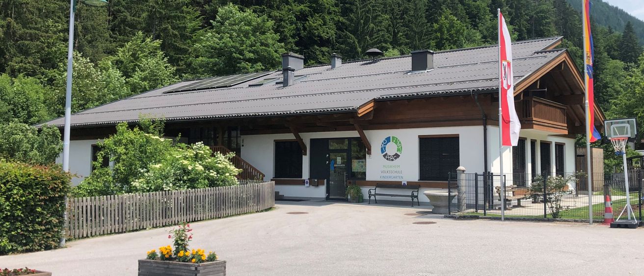 Ein Gebäude mit grauem Dach und Solarpanelen, umgeben von Bäumen und einem Zaun, mit Fahnen und einem Basketballkorb im Vordergrund.