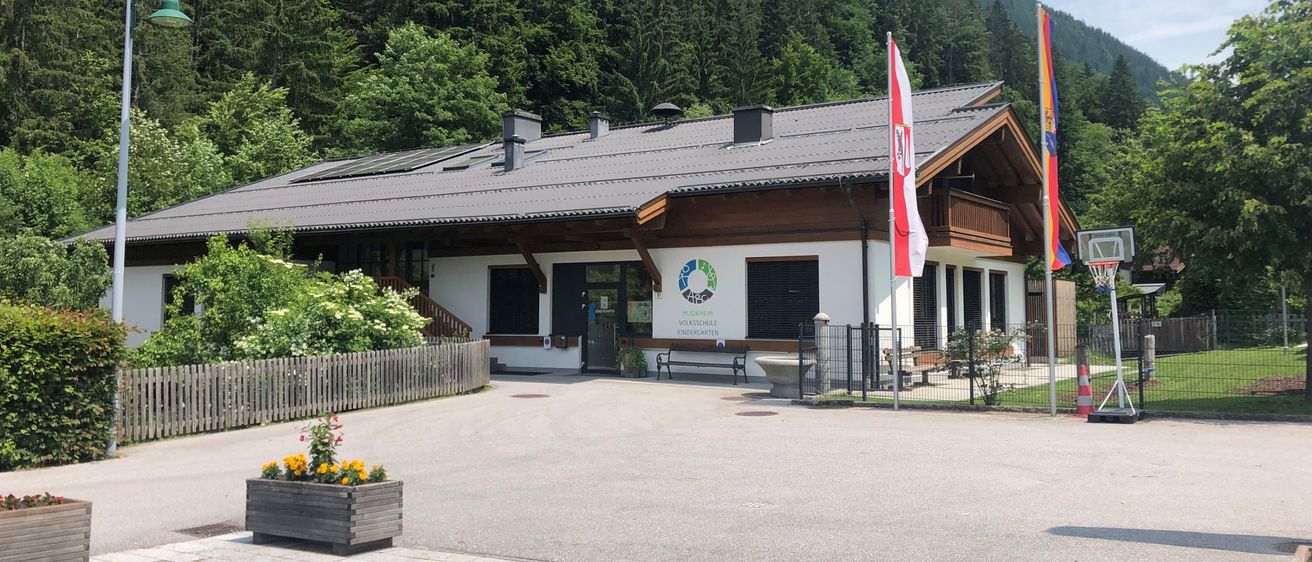 Ein Gebäude mit Solarpanelen auf dem Dach steht vor einem bewaldeten Hügel. Es verfügt über einen Holzzaun und zwei Flaggen, von denen eine die österreichische Flagge ist.