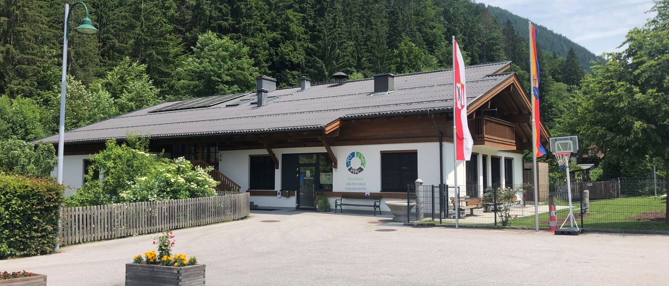Ein Gebäude mit Solarpanelen auf dem Dach steht vor einem bewaldeten Hügel. Es verfügt über einen Holzzaun und zwei Flaggen, von denen eine die österreichische Flagge ist.