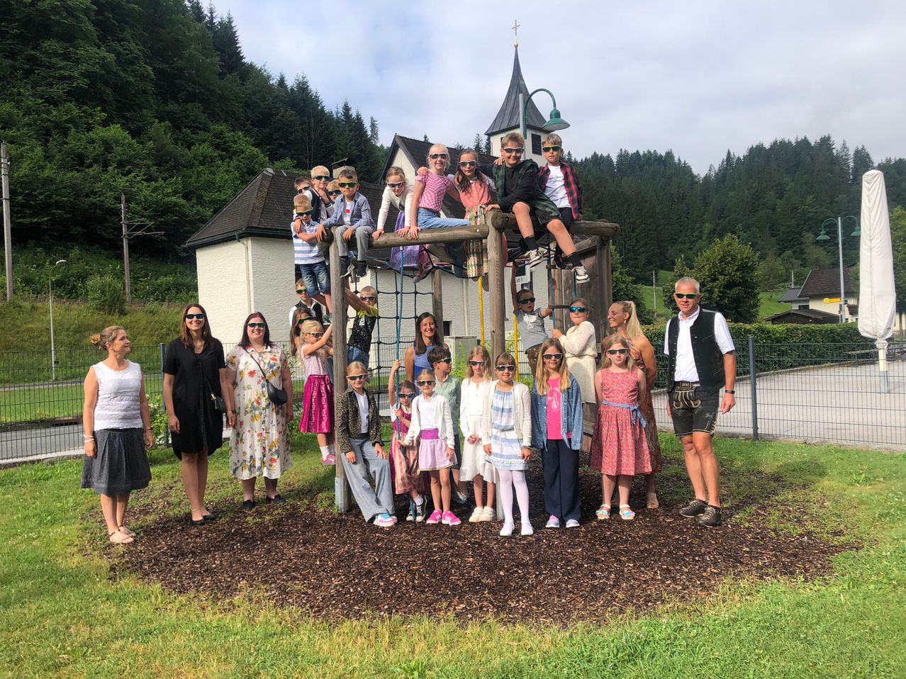Eine Gruppe von Menschen, einschließlich Erwachsener und Kindern, posiert für ein Foto in einem Park. Sie tragen Sonnenbrillen und einige sind auf einer Spielstruktur. Ein Haus und Bäume sind im Hintergrund.