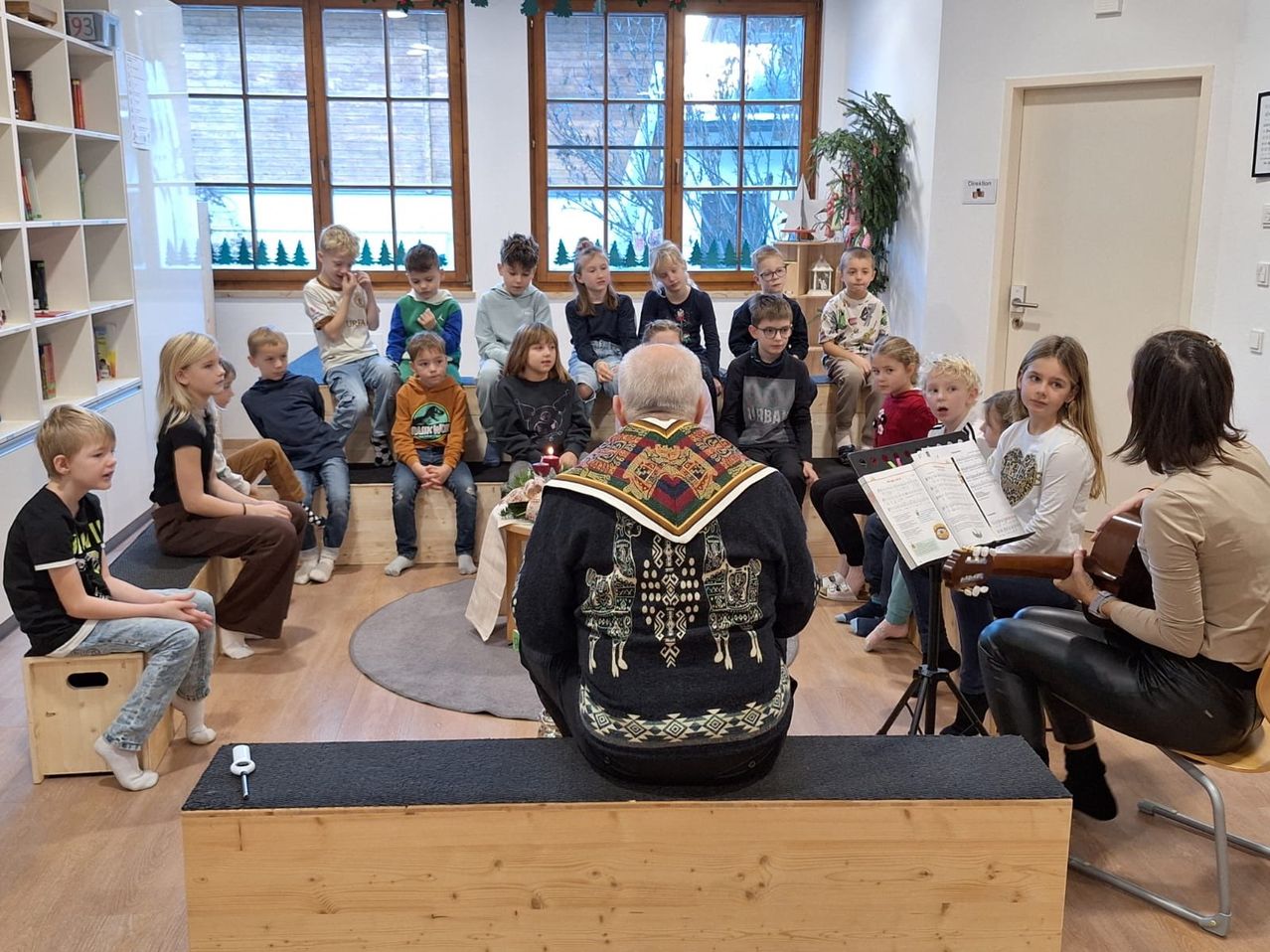 Eine Gruppe von Kindern ist in einem Raum mit Holzboden versammelt. Ein Erwachsener sitzt vor ihnen in einem dekorierten Pullover. Ein anderer Erwachsener spielt Gitarre. Einige Kinder halten Kerzen.
