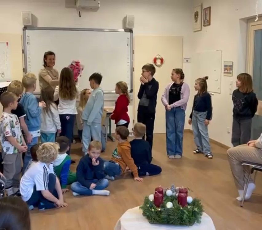 Ein Klassenzimmer voller Kinder ist um eine Lehrerin versammelt. Einige sitzen auf dem Boden, andere stehen. Die Lehrerin hält einen Blumenstrauß, und ein Tisch mit einem Kranz und Kerzen ist im Vordergrund.