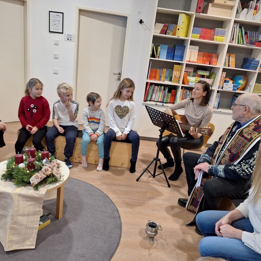 Eine Gruppe von Kindern und Erwachsenen sitzt um einen Tisch mit einer Gitarre, Kerzen und Blumen. Eine Frau spielt Gitarre, während ein Mann ein Buch liest. Ein Bücherregal voller Bücher steht hinter ihnen.