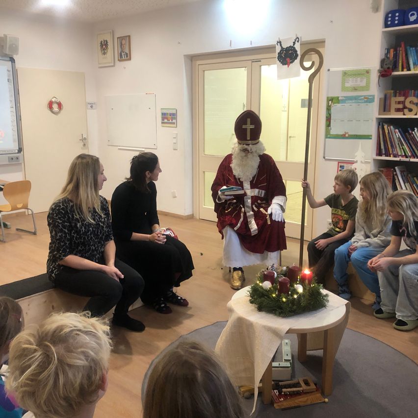 Eine Gruppe von Kindern und Erwachsenen sitzt um einen Tisch mit einem Kranz und Kerzen. Ein Mann in Santa Claus-Kostüm steht in der Mitte und hält einen Stab.