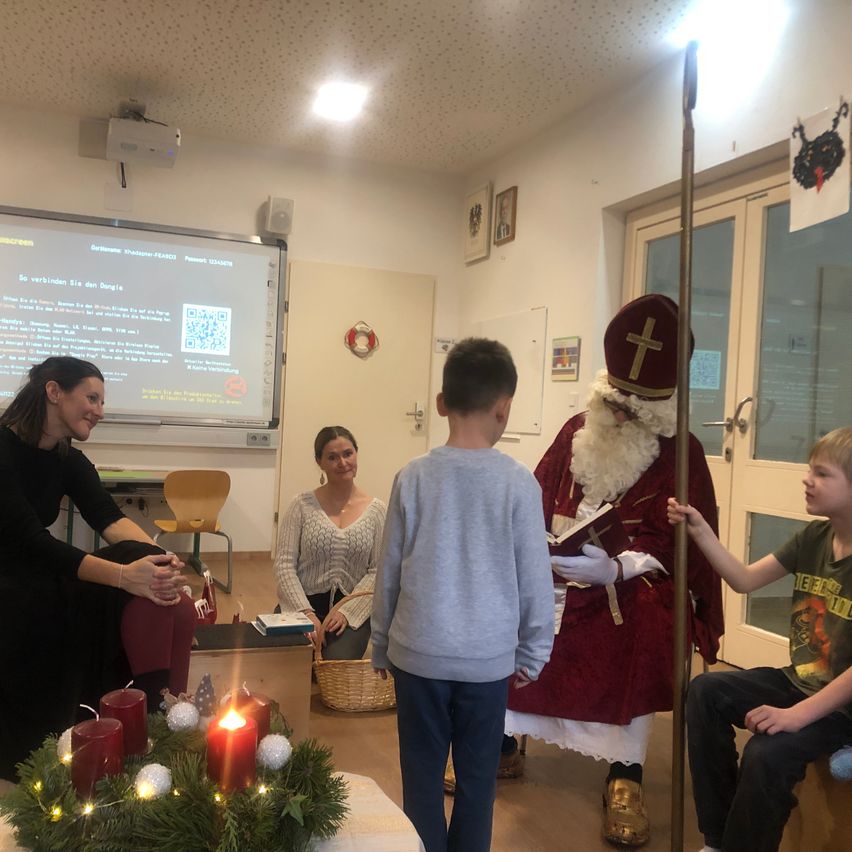 Eine Gruppe von Menschen versammelt sich in einem Raum. Ein Mann als Weihnachtsmann hält ein Buch und unterhält sich mit einem Jungen. Zwei Frauen sitzen auf dem Boden, eine trägt eine Halskette und hält ein Baby. Eine Beamerleinwand zeigt Informationen.