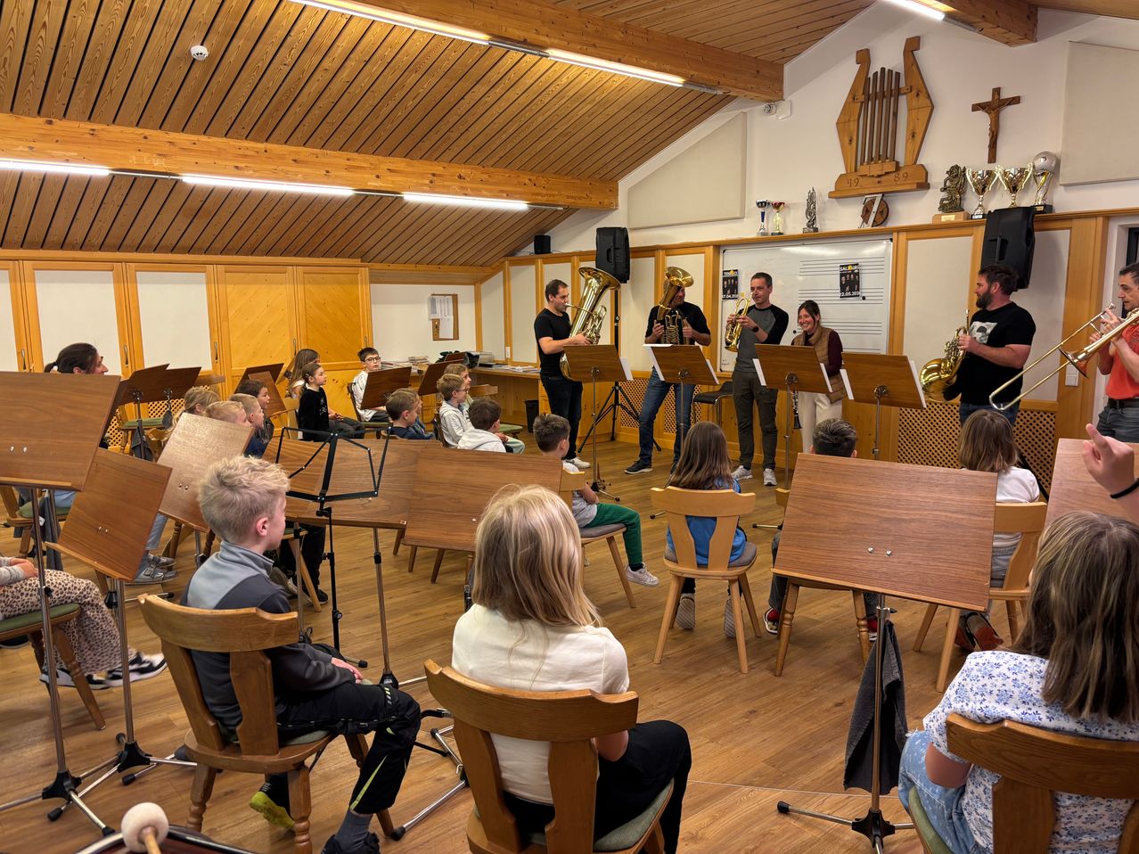 In einem Musikraum versammeln sich Erwachsene und Kinder. Einige Erwachsene spielen Blechblasinstrumente, während die Kinder sitzen und zuschauen. Der Raum ist mit Holzmöbeln, Notenständern und Trophäen ausgestattet.