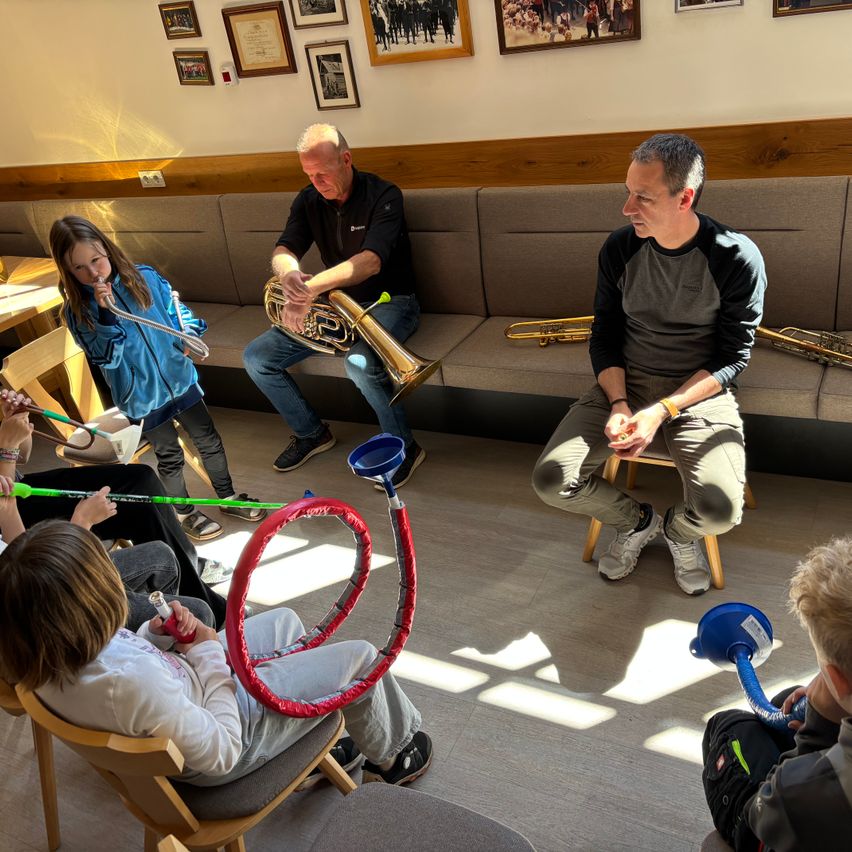 Eine Gruppe von Menschen in einem Raum, einige sitzen auf Stühlen und andere auf einer Couch, und üben mit Musikinstrumenten. Ein Mann sitzt auf einem Hocker und hält eine Trompete.
