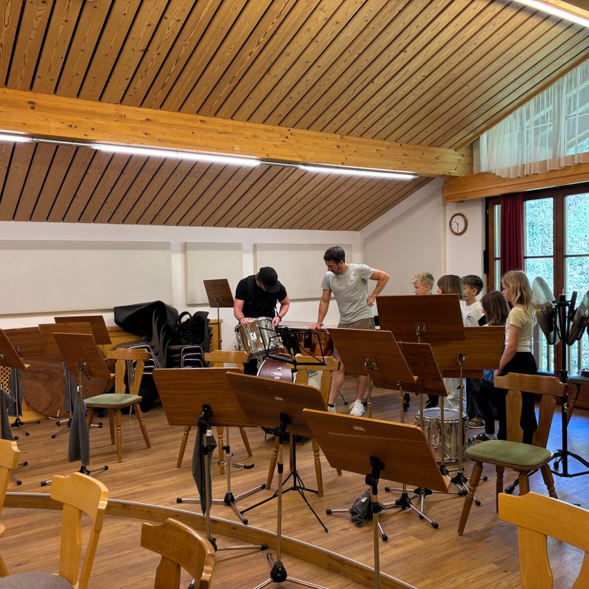 Zwei Männer spielen Trommeln in einem Musikraum, während Kinder zusehen. Der Raum hat Holzböden und -decken, und Musikinstrumente sind auf dem Boden aufgestellt.