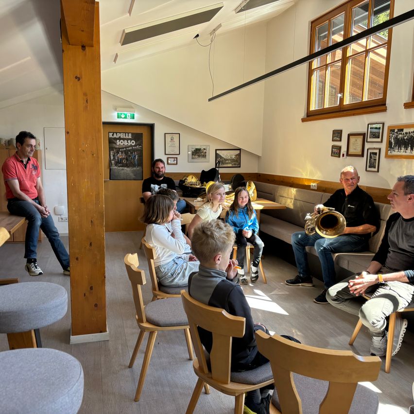 Eine Gruppe von Erwachsenen und Kindern sitzt in Stühlen um einen Raum herum. Ein Mann spielt eine Trompete. Ein Poster ist an der Wand hinter dem Mann.