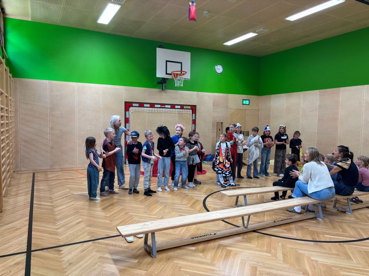 A group of children and adults gather in a gym, dressed in various costumes. A woman stands with them, and two wooden benches are nearby. A basketball hoop is mounted on the wall.