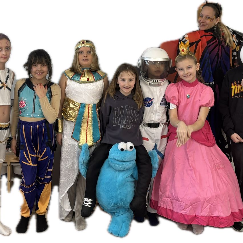 Eine Gruppe von Kindern in verschiedenen Kostümen posiert für ein Foto. Ein Kind ist als ägyptische Prinzessin gekleidet, ein anderes als Astronaut, und andere in verschiedenen Kostümen. Ein großes blaues Plüschtier ist ebenfalls vorhanden.