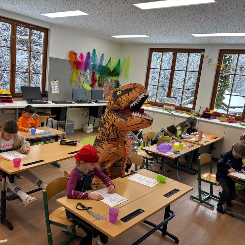 Ein Klassenzimmer mit Kindern, die Kunstaktivitäten durchführen; ein Kind ist als Dinosaurier verkleidet. Der Raum hat Holzböden, weiße Wände und große Fenster mit Schnee draußen.