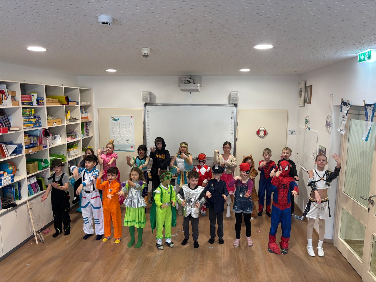 Eine Gruppe von Kindern in verschiedenen Kostümen, darunter Superhelden, Astronauten und andere Charaktere, posiert für ein Foto in einem Klassenzimmer.