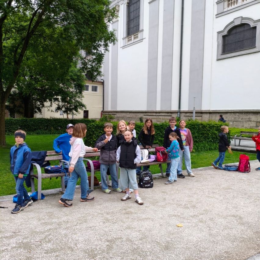 Eine Gruppe von Kindern mit Rucksäcken versammelt sich vor einer Kirche, einige stehen, andere sitzen auf einer Bank, mit Büschen und einem Weg im Vordergrund.