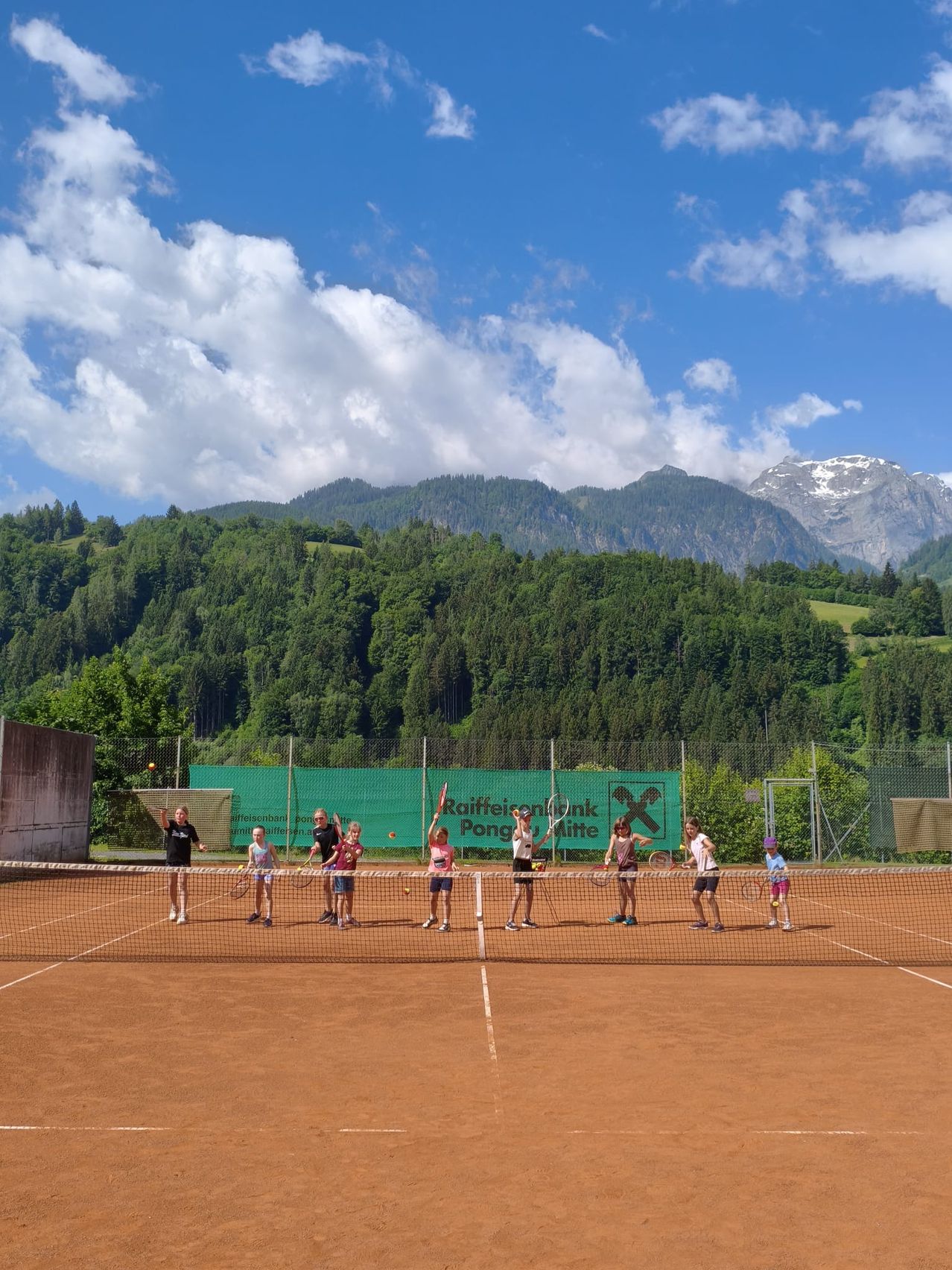 Eine Gruppe von Menschen spielt Tennis auf einem Tennisplatz mit Bergen im Hintergrund.