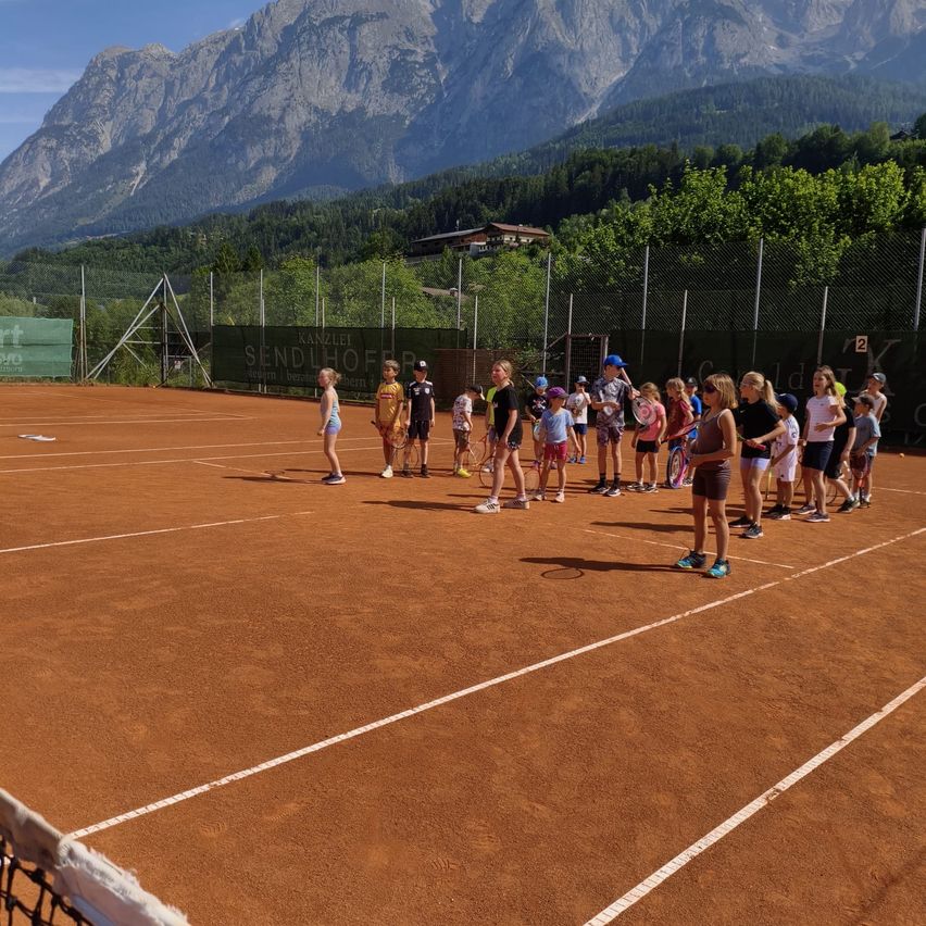 Kinder in Sportkleidung stehen auf einem Tennisplatz und halten Tennisschläger. Eine Bergkette und Grünpflanzen sind im Hintergrund.