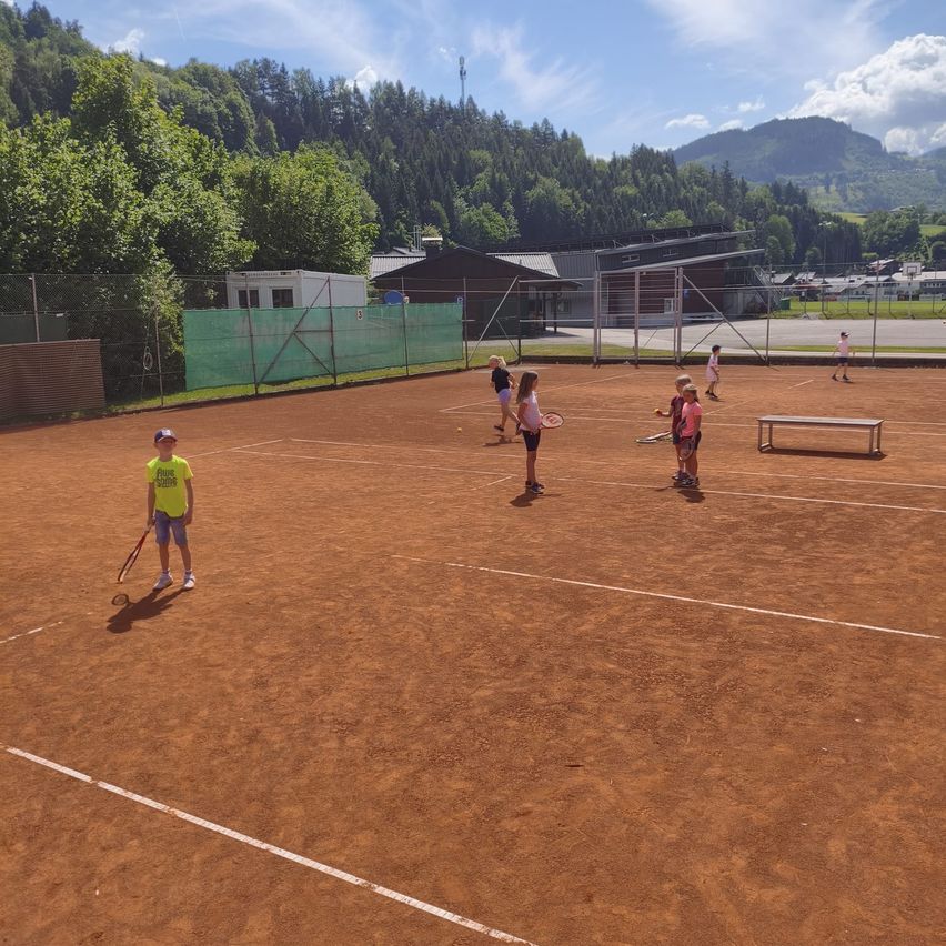 Kinder spielen Tennis auf einem Sandplatz, mit Bergen und Bäumen im Hintergrund. Es gibt mehrere Spieler, einige mit Mützen, die Tennisschläger halten.