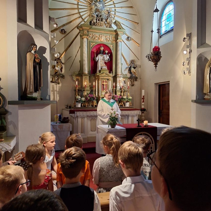 A priest stands at an altar in a church, surrounded by children listening attentively. The altar is adorned with flowers and candles, and statues and religious symbols are visible in the background.