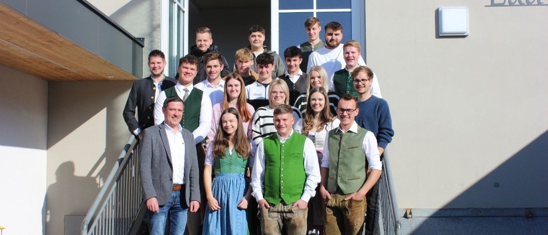 Eine Gruppe von Menschen, in verschiedenen Kleidungsstücken gekleidet, steht auf den Stufen eines Gebäudes, möglicherweise für ein Gruppenfoto. Das Gebäude hat eine Glastür und eine Wand mit dem Namen Ludwig.