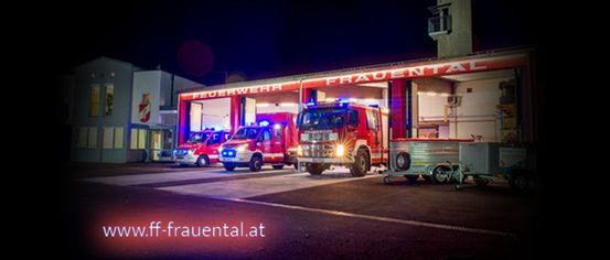 Three fire trucks are parked in the Frauental fire station at night. The lights are on, and the station has a red roof.