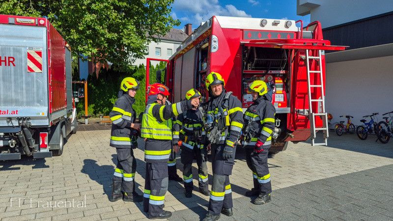 Eine Gruppe Feuerwehrleute in Uniform, darunter ein Mann, der zeigt, steht vor einem Feuerwehrauto. Dahinter befindet sich ein Gebäude mit einem Baum und Fahrrädern.