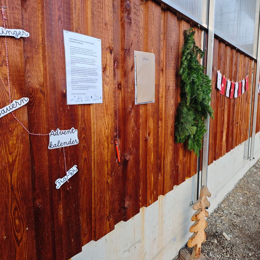 Eine Holzwand mit Dekorationen und Texten, die angeheftet sind. 'Adventskalender' und 'Frau Di' stehen auf hängenden Papieren. Eine grüne Girlande hängt an der Wand, und rechts ist eine Fahne angebracht.