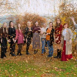 Eine Gruppe von Frauen in Herbstkleidung steht auf einem Feld mit umherfliegenden Blättern und lächelt für ein Foto.