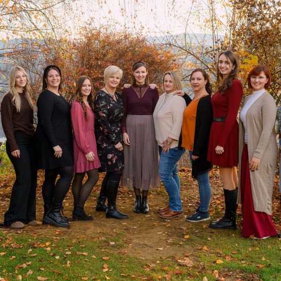Eine Gruppe von Frauen, alle festlich gekleidet, posiert für ein Foto im Freien mit Herbstlaub am Boden.