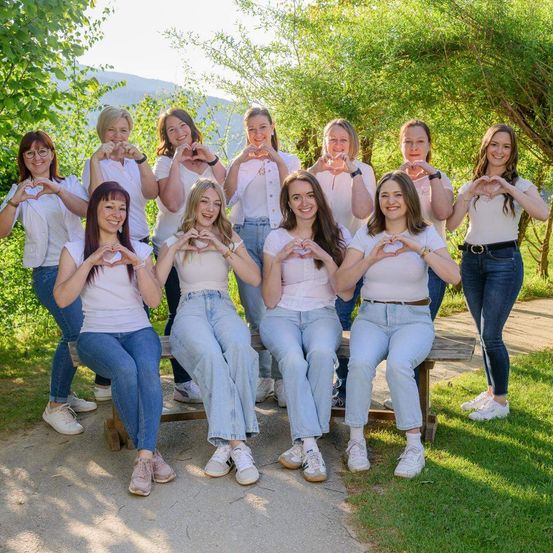 Eine Gruppe von Frauen, angezogen in weißen Shirts und Jeans, posiert für ein Foto und macht Herzformen mit ihren Händen, auf einer Bank umgeben von Bäumen.