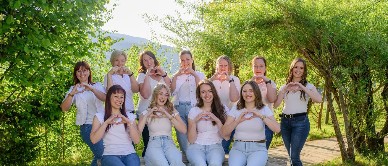 Eine Gruppe von Frauen in passenden Outfits posiert mit herzförmigen Handgesten in einem Park, umgeben von Bäumen und Gras.