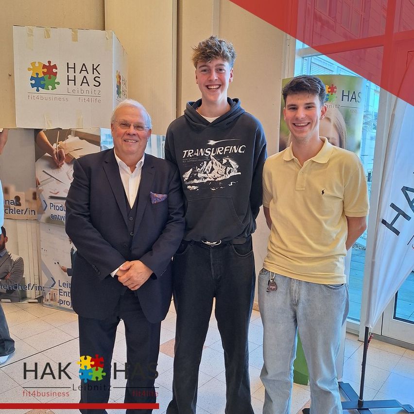 Drei Personen stehen in einer Lobby zusammen, zwei Männer lächeln und einer trägt eine Brille. Hinter ihnen befinden sich Banner mit Logos.